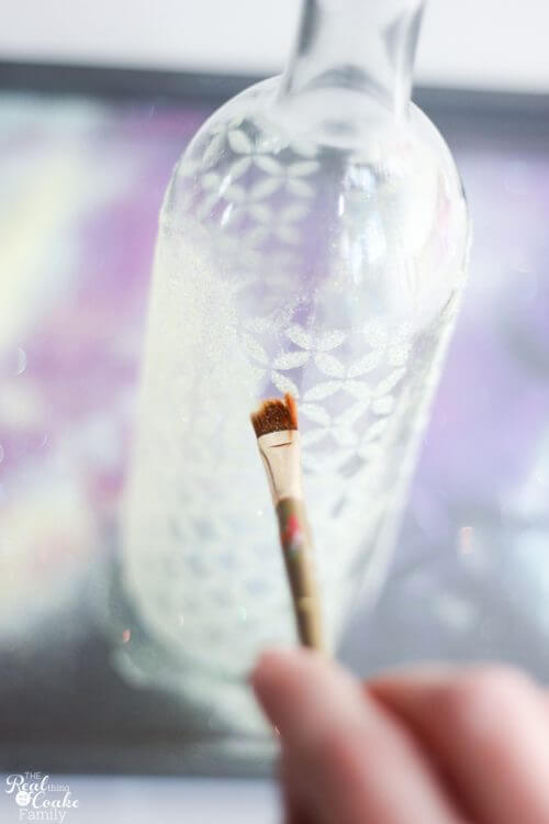 Wine bottle crafts are so fun. These are beautiful DIY vases made with Mod Podge, stencils and glitter. Great for Spring home decor or change the colors for anytime of year. Love these! #WineBottleCrafts #WineBottle #Crafts #ModPodge #Glitter paintbrush gently removing extra glitter from wine bottle